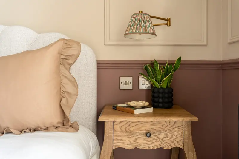 Bedside detail with a ruffled linen cushion, carved wooden side table and plum panelled wall