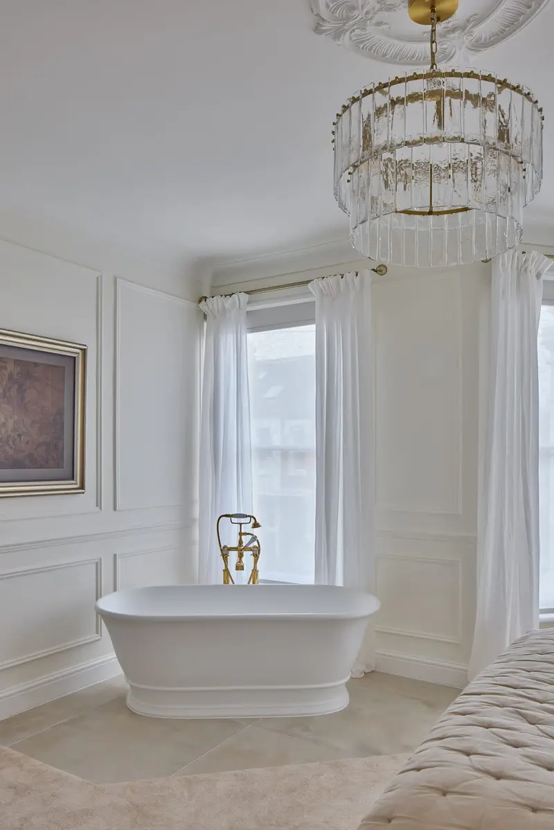 Freestanding roll-top bath beneath a chandelier in a bay window